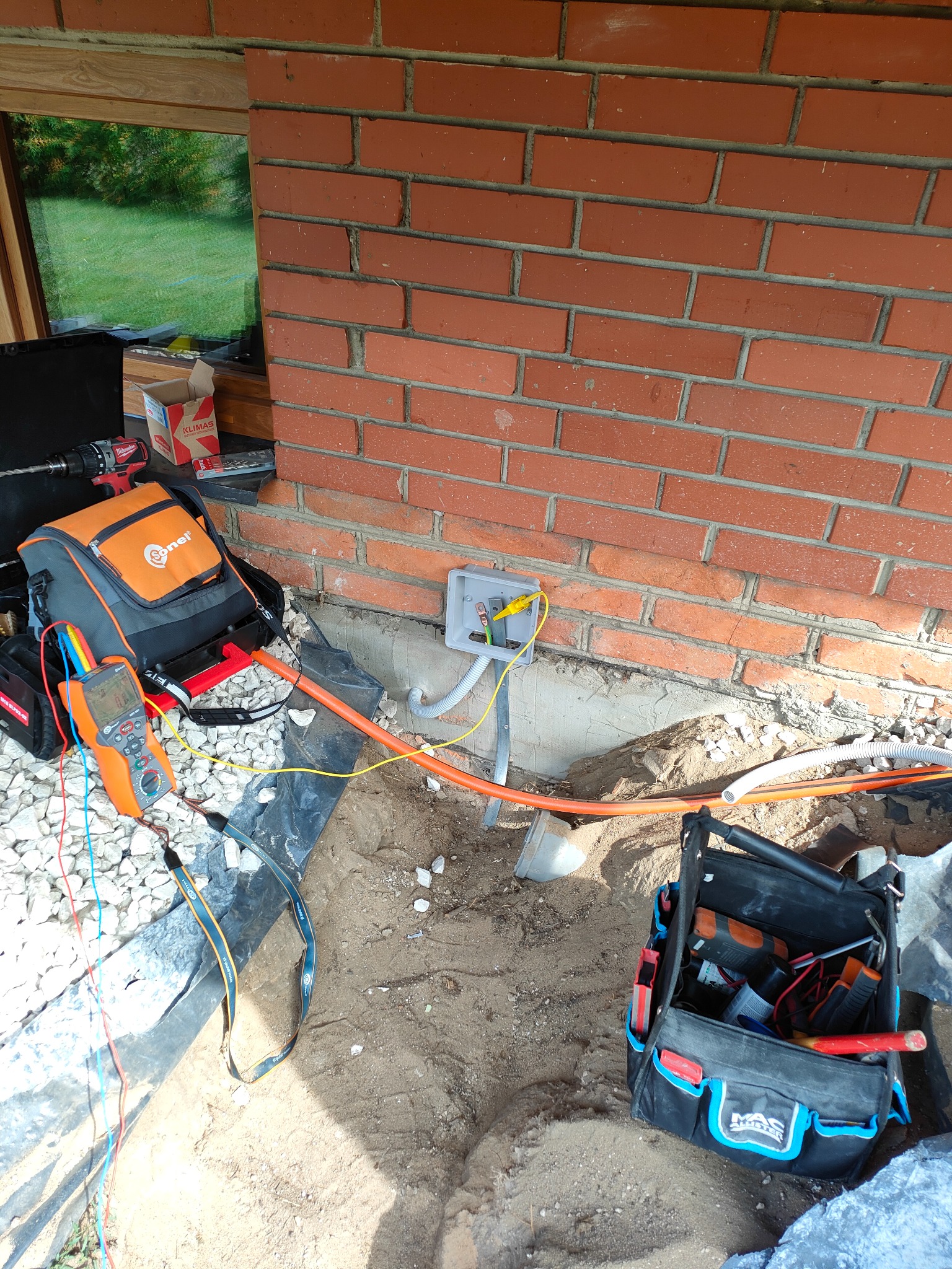 Prace ziemne i podłączenie kabli w puszce elektrycznej na zewnątrz budynku w Radomiu, widoczny miernik, torba narzędziowa i przewody.