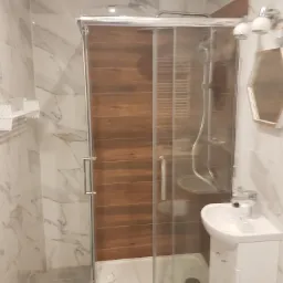 Nowoczesna łazienka z kabiną prysznicową z przesuwnymi drzwiami, wykończona płytkami imitującymi marmur i drewno, mała umywalka z szafką, sześciokątne lustro i biała suszarka na ścianie.