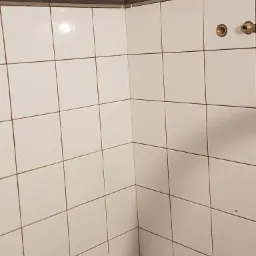 Ściana w rogu wyłożona białymi, kwadratowymi płytkami ceramicznymi z widocznymi elementami mocującymi i przyłączami wodnymi.