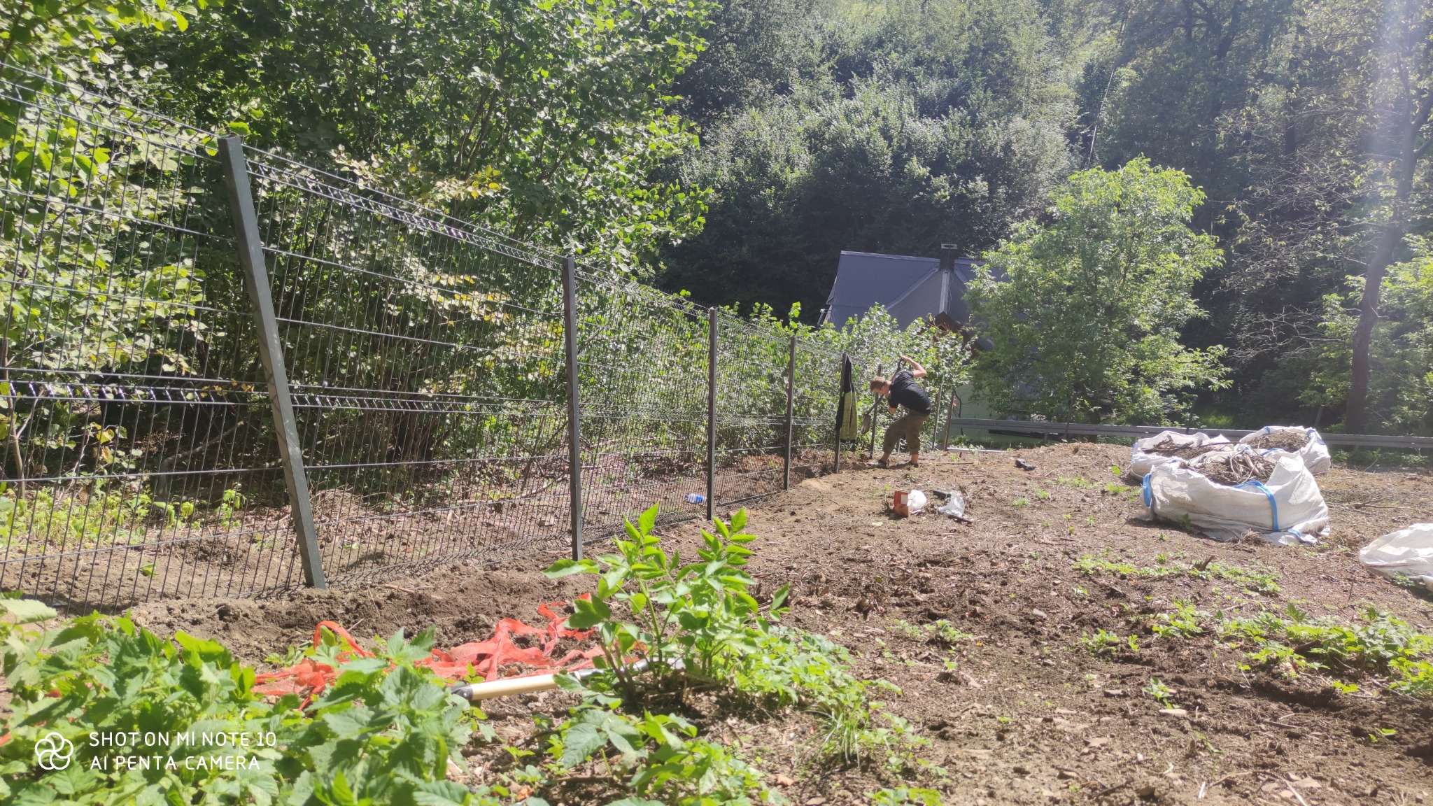 Montaż szarego ogrodzenia panelowego na nierównym terenie leśnym, widoczna osoba kopiąca dół pod słupek i worki z ziemią w tle.