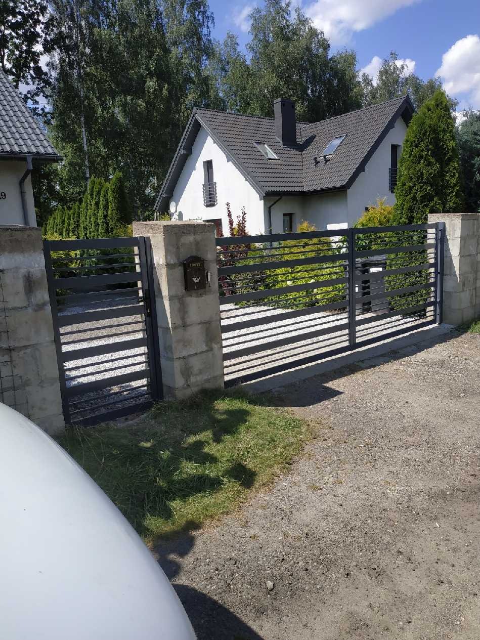 Nowoczesna, pozioma brama wjazdowa z szarych, płaskich elementów, osadzona na betonowych słupkach z zamontowaną skrzynką na listy, na tle domu z ciemną dachówką i zielenią ogrodu.