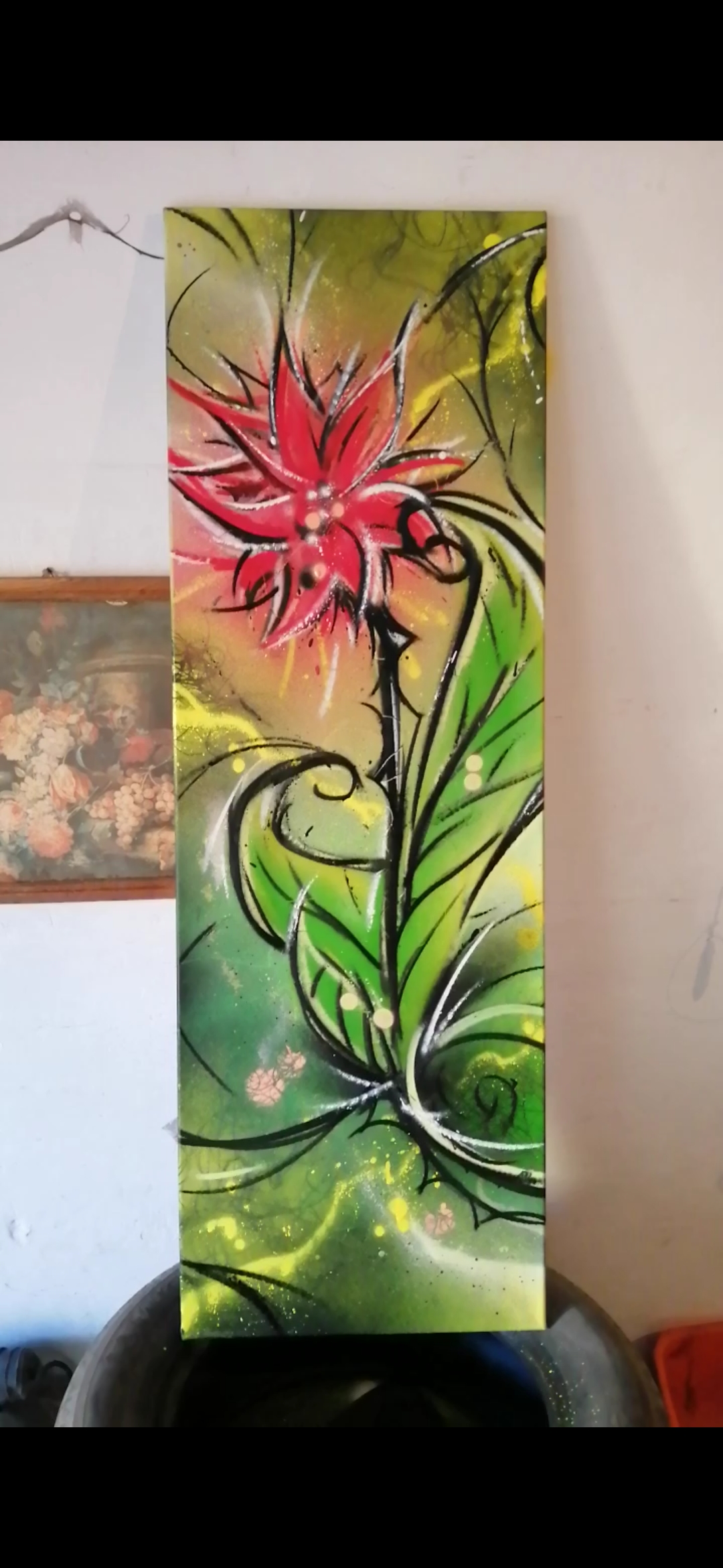 Wertykalny obraz przedstawiający stylizowany czerwony kwiat z zielonymi liśćmi na rozmytym, zielono-żółtym tle z czarnymi konturami, umieszczony obok obrazu w ramie wiszącego na ścianie.