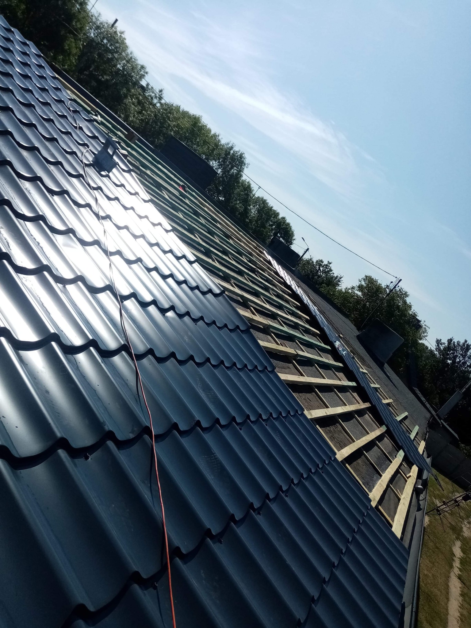 Częściowo pokryty dach z blachy trapezowej w kolorze grafitowym, widoczna konstrukcja drewniana i fragmenty starego pokrycia, ujęcie pod kątem w słoneczny dzień.