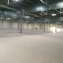 Jasna, gładka posadzka w hali przemysłowej z metalową konstrukcją dachu i filarami. Widoczne światło dzienne wpadające przez świetliki dachowe i otwarte drzwi.