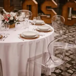 Elegancko nakryty okrągły stół z białym obrusem, przezroczystymi krzesłami i kwiatową dekoracją, na tle dużych, podświetlanych liter dekoracyjnych i białego namiotu.