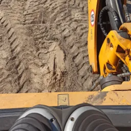 Widok z perspektywy operatora żółtej koparko-ładowarki JCB podczas niwelacji terenu piaszczystego gruntu, widoczne ślady opon i świeżo wyrównana powierzchnia.