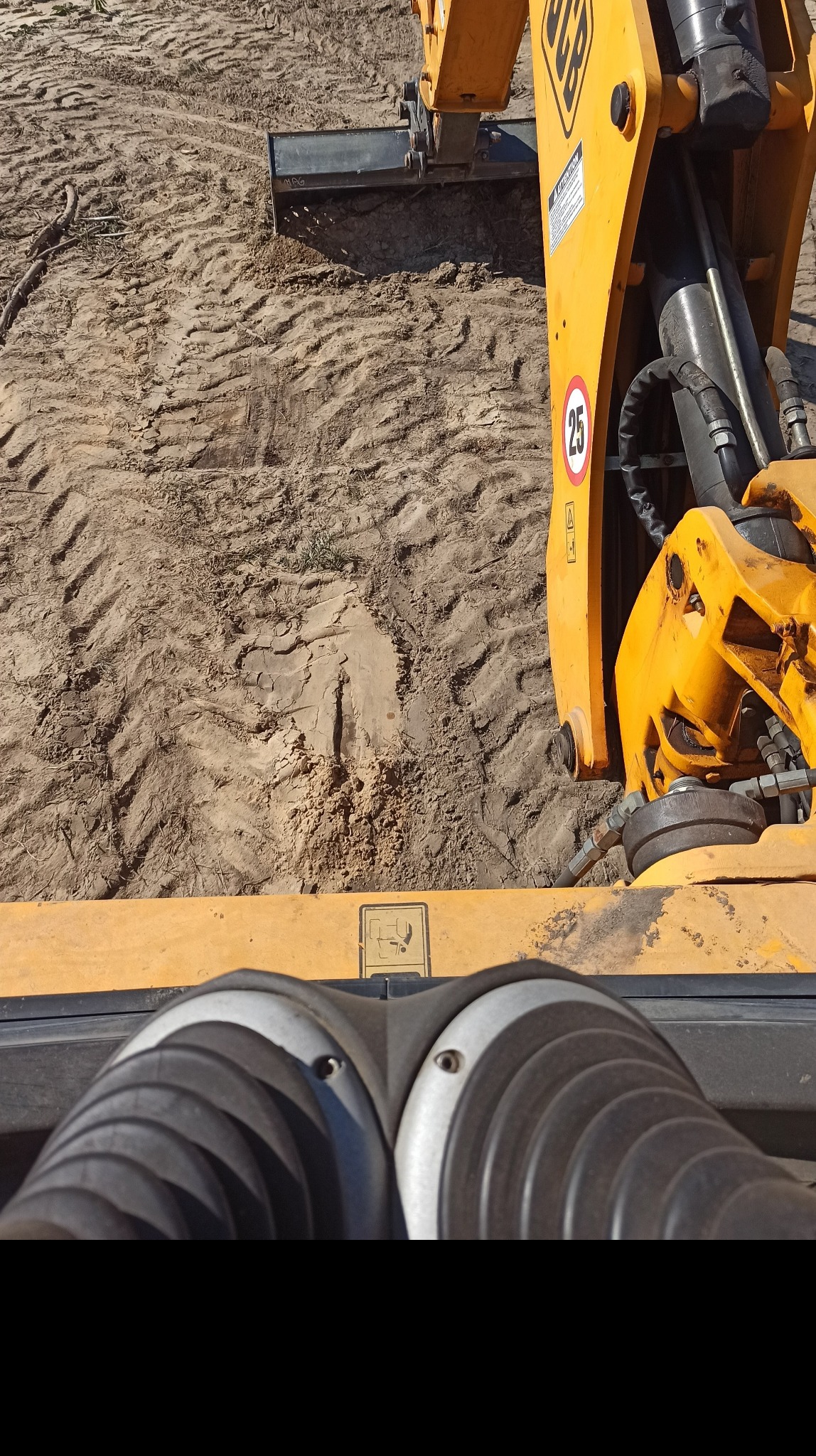 Widok z perspektywy operatora żółtej koparko-ładowarki JCB podczas niwelacji terenu piaszczystego gruntu, widoczne ślady opon i świeżo wyrównana powierzchnia.