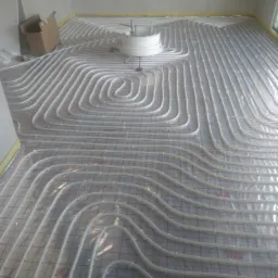 Rozłożone białe rury ogrzewania podłogowego na folii z czerwoną siatką, widoczny zwój rur i karton w rogu pomieszczenia.