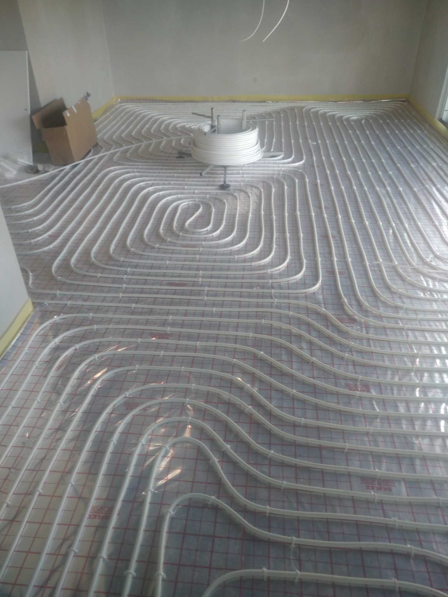 Rozłożone białe rury ogrzewania podłogowego na folii z czerwoną siatką, widoczny zwój rur i karton w rogu pomieszczenia.