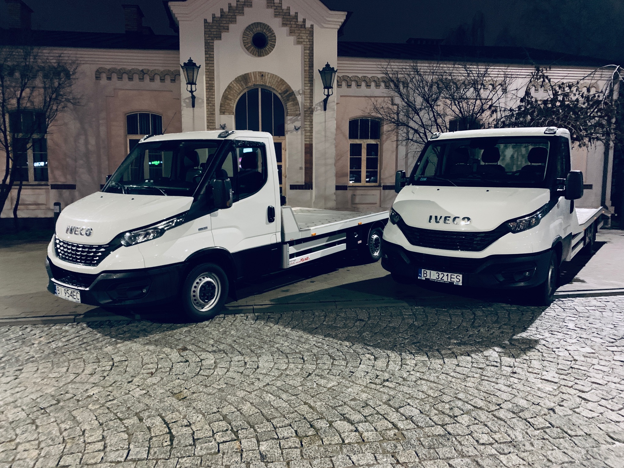 Dwa białe samochody ciężarowe Iveco z platformą, zaparkowane na brukowanej ulicy przed budynkiem z cegły w stylu retro, nocne oświetlenie.
