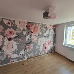 Pokój z fototapetą w kwiaty, panele podłogowe przygotowane do montażu, widoczny grzejnik i nowoczesna lampa sufitowa.