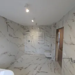 Wykończona łazienka z białymi płytkami imitującymi marmur, grzejnikiem drabinkowym, toaletą podwieszaną i deszczownicą.