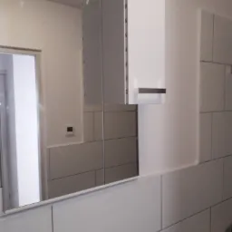 Nowoczesna łazienka z białymi płytkami i szafką z lustrem, widoczny fragment kabiny prysznicowej w odbiciu lustra.