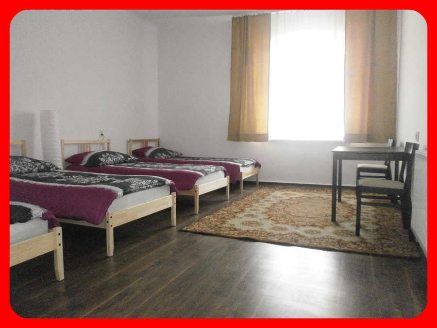 Prosty pokój z trzema łóżkami pojedynczymi, nakrytymi bordowymi narzutami i wzorzystą pościelą, stolik z krzesłami przy oknie z jasnymi zasłonami, dywan na podłodze imitującej drewno.