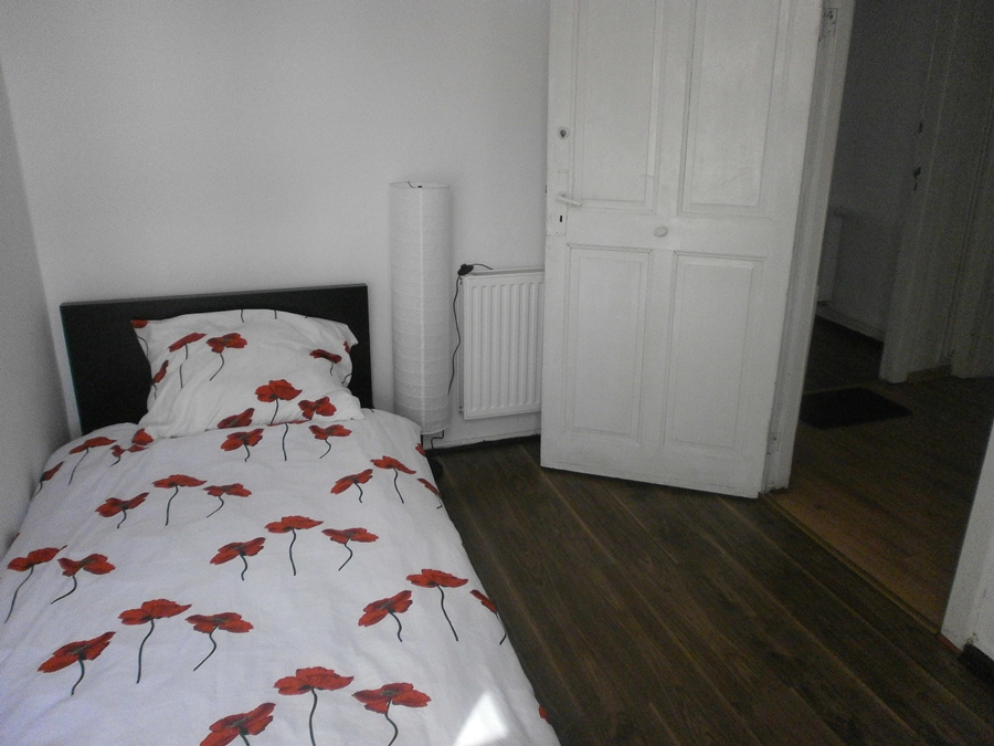 Pokój z pojedynczym łóżkiem z pościelą w czerwone maki, białą lampą stojącą i otwartymi, białymi drzwiami.