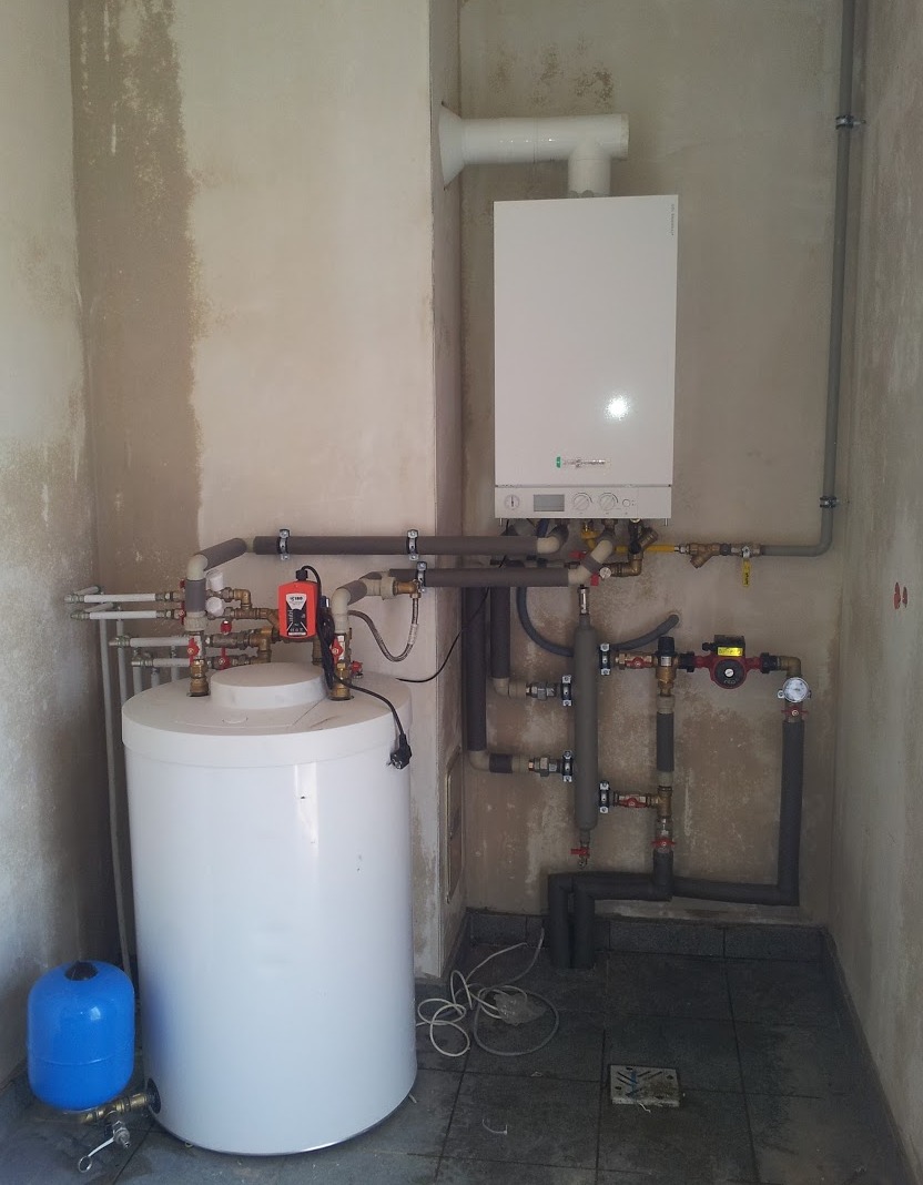 Biały piec CO wiszący na ścianie, z podłączonym zasobnikiem ciepłej wody i niebieskim zbiornikiem wyrównawczym, liczne rury i zawory, surowe ściany w tle.