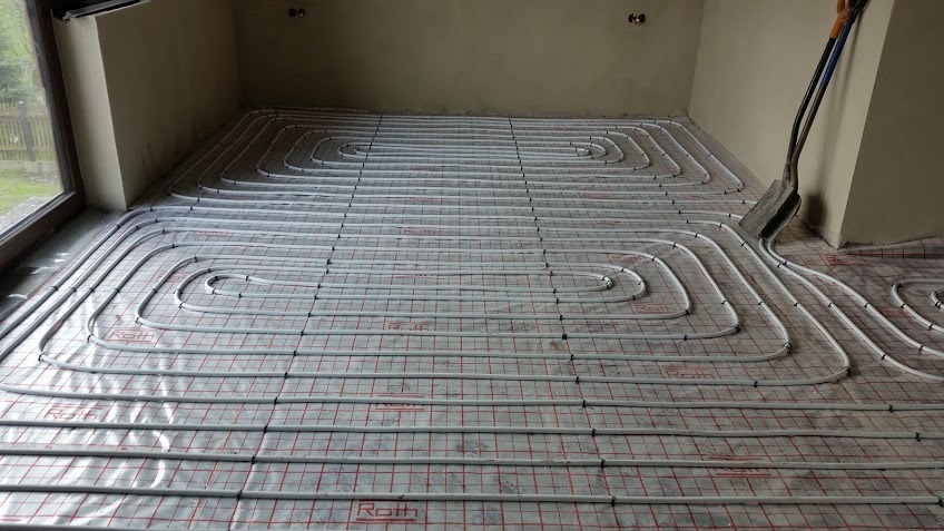 Rozłożone rury ogrzewania podłogowego marki Roth na folii z siatką, widoczne narzędzia opierające się o ścianę.