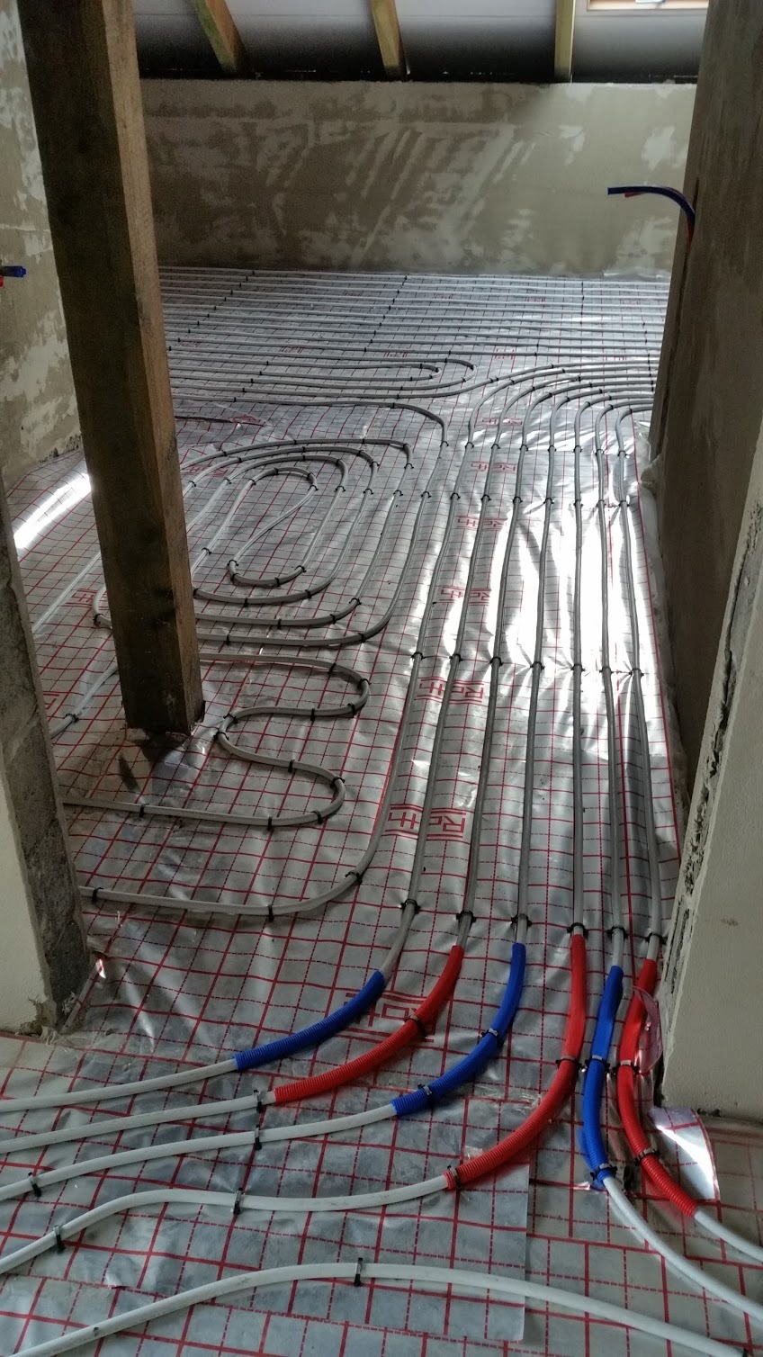 Montaż ogrzewania podłogowego w surowym stanie, rury grzewcze ułożone na folii z siatką, z czerwono-niebieskimi osłonami na końcach przy ścianie, widoczny drewniany słup konstrukcyjny.