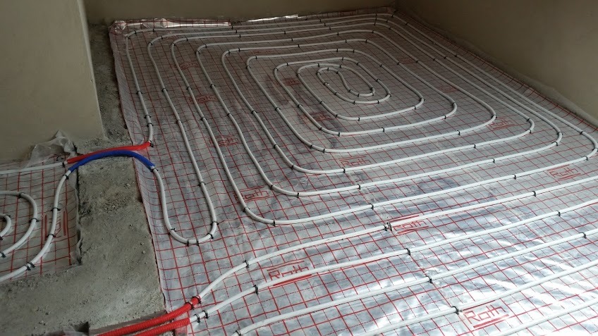 Białe rury ogrzewania podłogowego Roth, ułożone spiralnie na folii z czerwoną kratą, w surowym, betonowym pomieszczeniu w trakcie instalacji.
