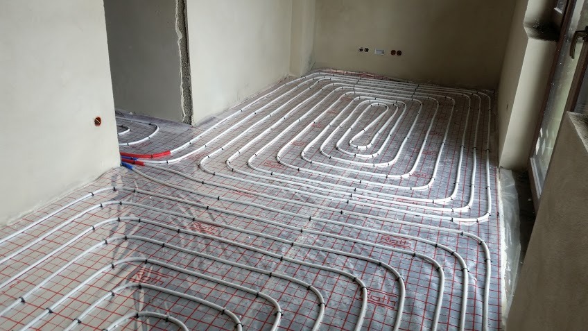 Białe rury ogrzewania podłogowego ułożone spiralnie na podkładzie z siatką w surowym wnętrzu, widoczne logo Roth na rurach.