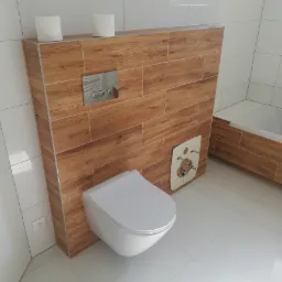 Nowoczesna toaleta podwieszana, obudowana płytkami imitującymi drewno, z widoczną instalacją wodną i przyciskiem spłukującym, na tle białych płytek ceramicznych na ścianach i podłodze.