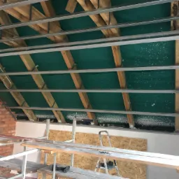 Szkielet metalowy podwieszanego sufitu na drewnianej konstrukcji dachu, zielona folia izolacyjna widoczna powyżej, ślady pianki montażowej, rusztowanie i drabina w pomieszczeniu z ceglaną ścianą.