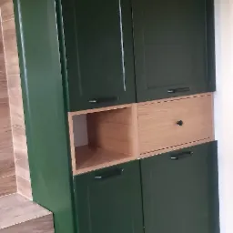 Wysoka, zielona szafka z drewnianymi elementami w kolorze naturalnego drewna, umieszczona w rogu pomieszczenia z jasnymi płytkami na podłodze i ścianie, obok drewnianego blatu. Szafka ma cztery...