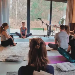 Grupa osób siedzi na matach w przestronnym pokoju z dużym oknem, wykonując ćwiczenia relaksacyjne; widoczny mikrofon na statywie.