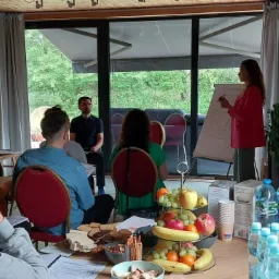 Grupa osób uczestniczy w warsztatach z flipchartem, gdzie kobieta w różowej marynarce prezentuje rysunek koła ze strzałkami, na stole przekąski, owoce i woda.