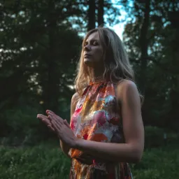 Kobieta z blond włosami w kwiecistej sukience, z zamkniętymi oczami, trzyma dłonie przed sobą w lesie, prawdopodobnie podczas medytacji.