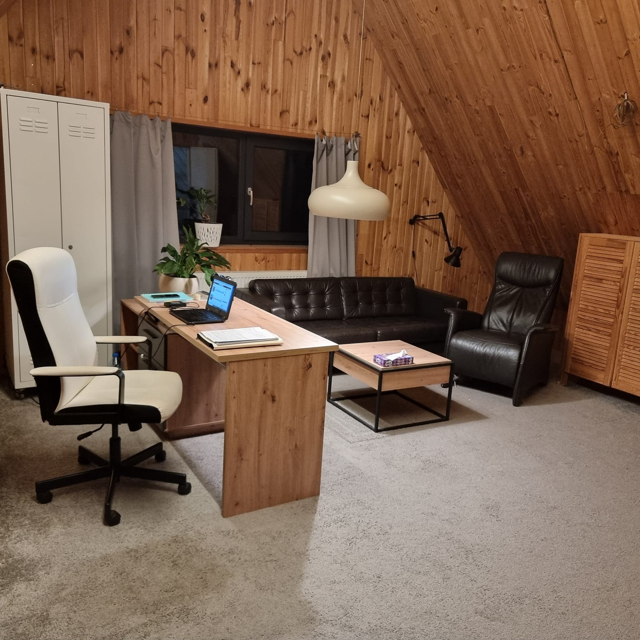 Pokój na poddaszu z drewnianą boazerią, biurkiem z laptopem, sofą i fotelem ze skóry, oświetlony wiszącą lampą.