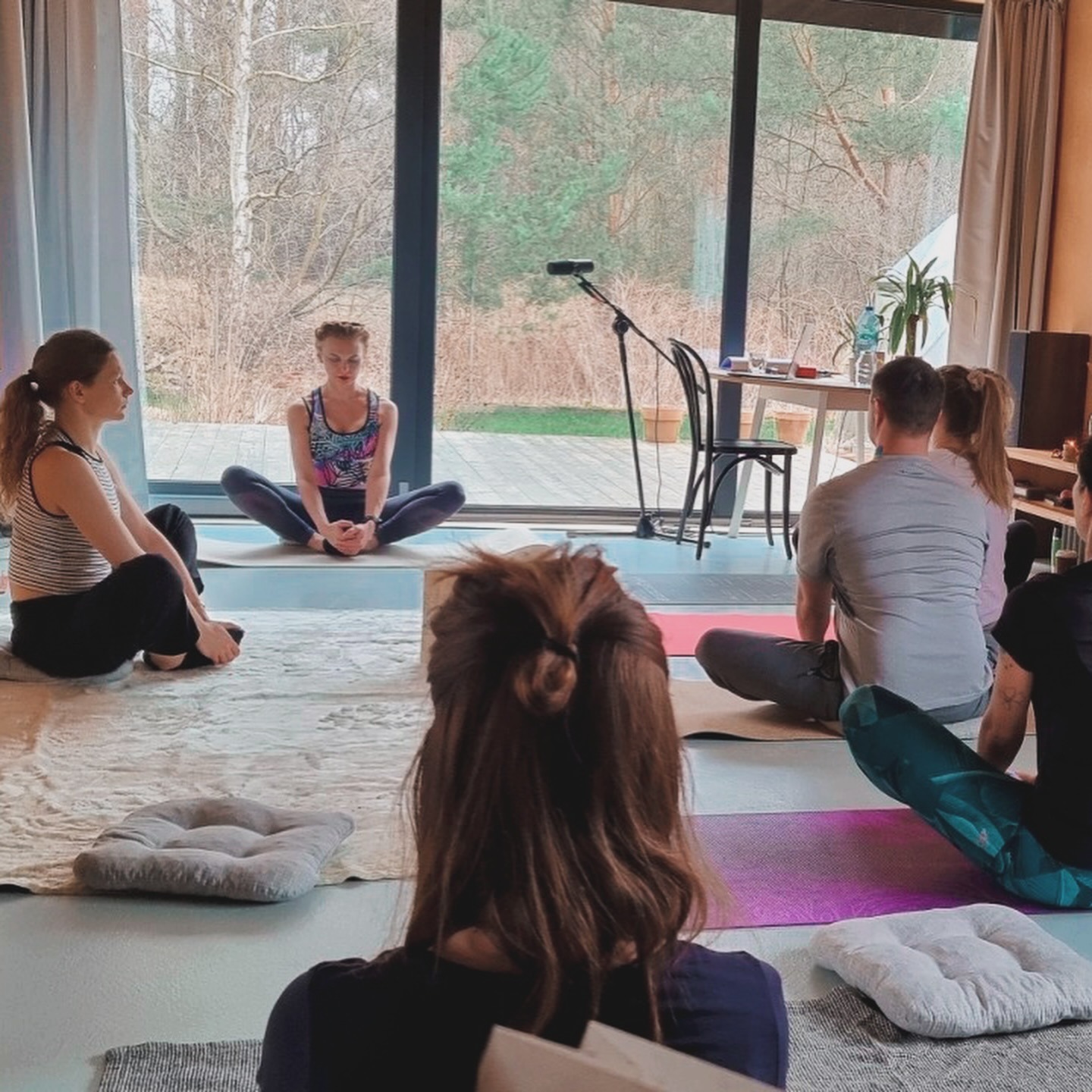 Grupa osób siedzi na matach w przestronnym pokoju z dużym oknem, wykonując ćwiczenia relaksacyjne; widoczny mikrofon na statywie.
