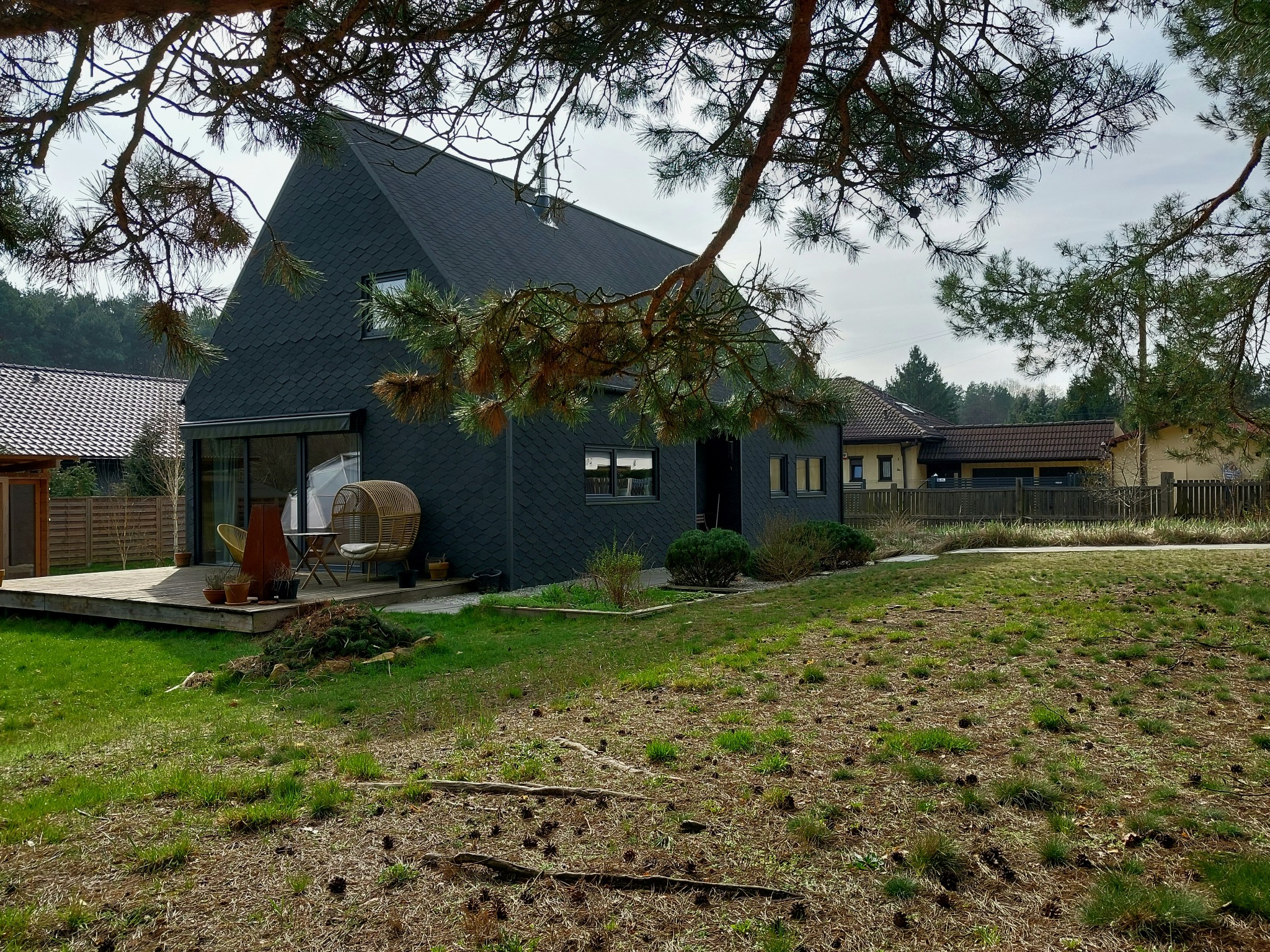 Nowoczesny dom z czarną elewacją i dachem z łupka, widok z ogrodu z drewnianym tarasem i ażurowymi meblami, częściowo przysłonięty gałęziami sosny.