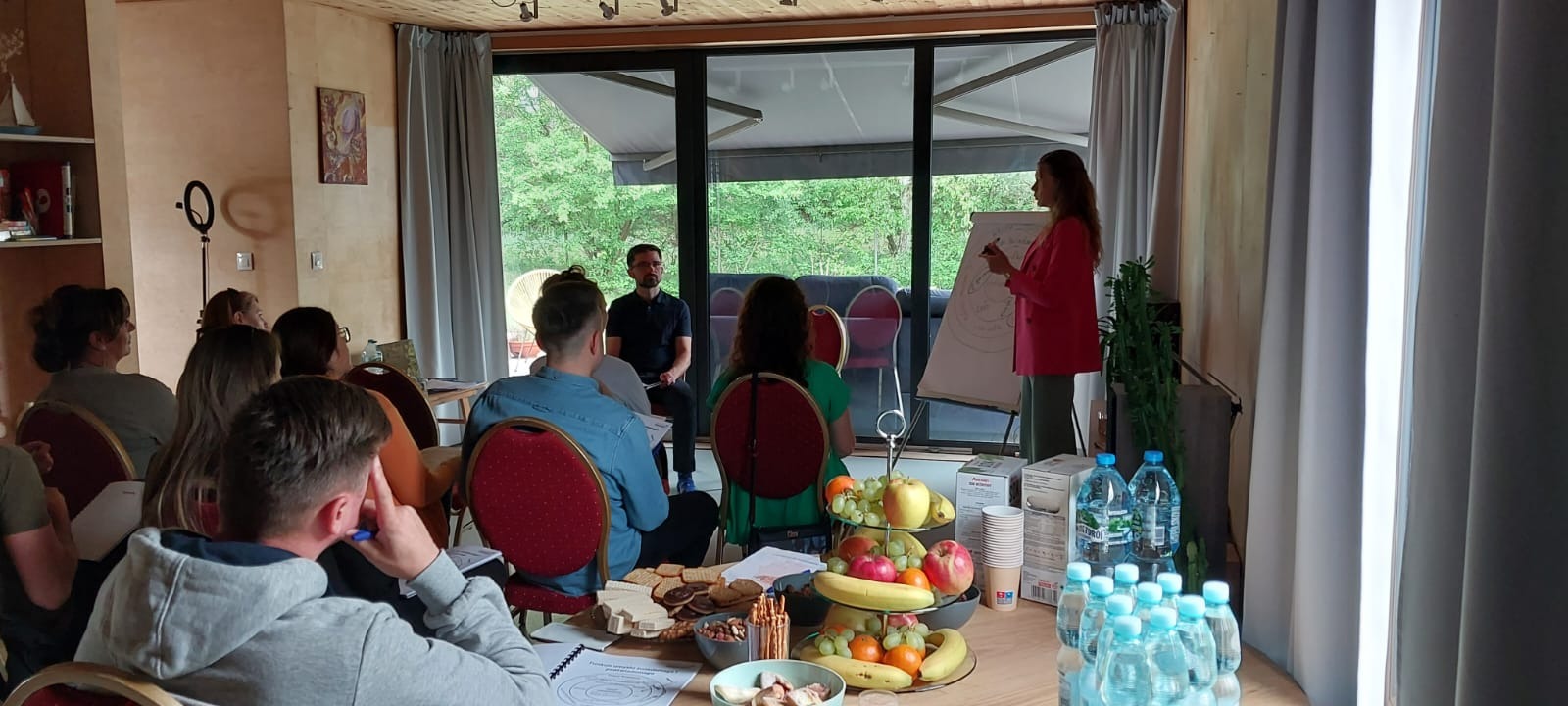 Grupa osób uczestniczy w warsztatach z flipchartem, gdzie kobieta w różowej marynarce prezentuje rysunek koła ze strzałkami, na stole przekąski, owoce i woda.