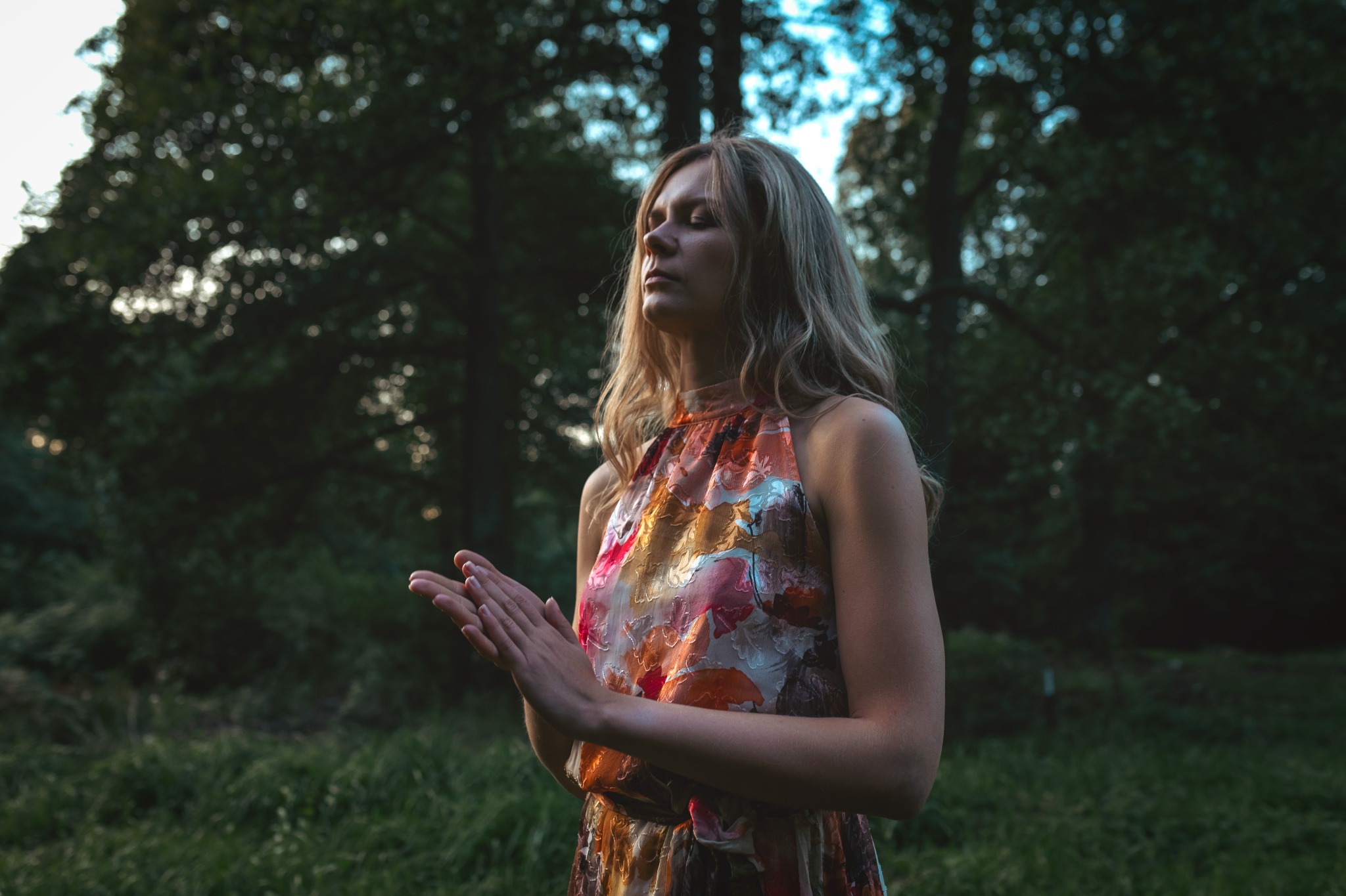 Kobieta z blond włosami w kwiecistej sukience, z zamkniętymi oczami, trzyma dłonie przed sobą w lesie, prawdopodobnie podczas medytacji.