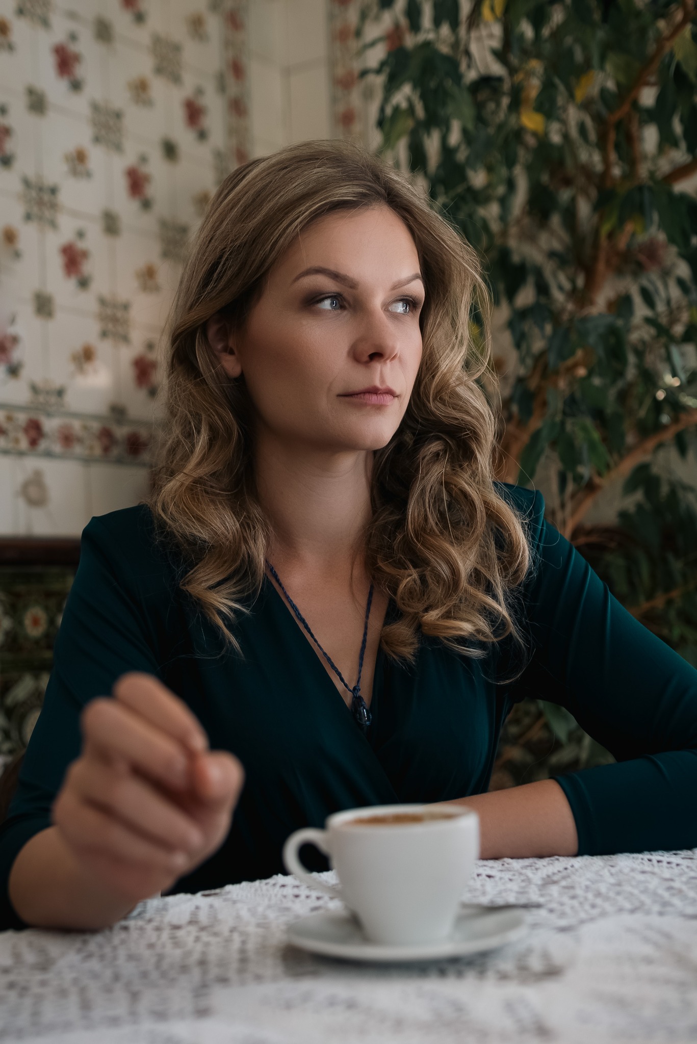 Portret kobiety z blond włosami, ubrana w zieloną sukienkę, siedzącej przy stole z filiżanką kawy, spoglądającej w bok, z delikatnym uśmiechem na tle wzorzystej tapety i rośliny.