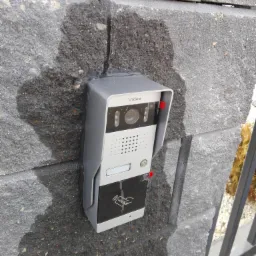 Szary wideodomofon marki Vidos zamontowany na szarym, betonowym murze ogrodzeniowym, z widocznymi śladami po montażu i uszczelnieniu, obok fragment metalowego ogrodzenia.