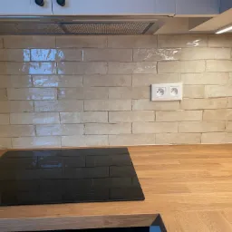 Beżowe, błyszczące płytki ułożone w cegiełkę na ścianie nad blatem kuchennym z zamontowaną płytą indukcyjną; widoczne gniazdko elektryczne i okap.