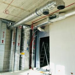 Daniel Majewski - Wnętrze w trakcie montażu systemu wentylacyjnego: widoczne metalowe rury wentylacyjne pod sufitem, pionowe kanały owinięte folią izolacyjną, drabina oparta o ścianę w przejściu, oraz przewody...