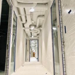 Daniel Majewski - Długi korytarz w budynku komercyjnym w trakcie prac wykończeniowych, widoczne białe rury wentylacyjne pod sufitem, zabezpieczone folią okna i ściany, niebieska taśma malarska.