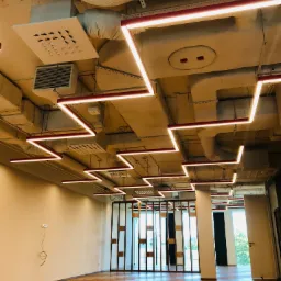 Wnętrze biurowe z widocznymi elementami wentylacji i klimatyzacji pod sufitem, w tym kanały wentylacyjne, kratki nawiewne i system oświetlenia liniowego LED w kształcie zygzaka.