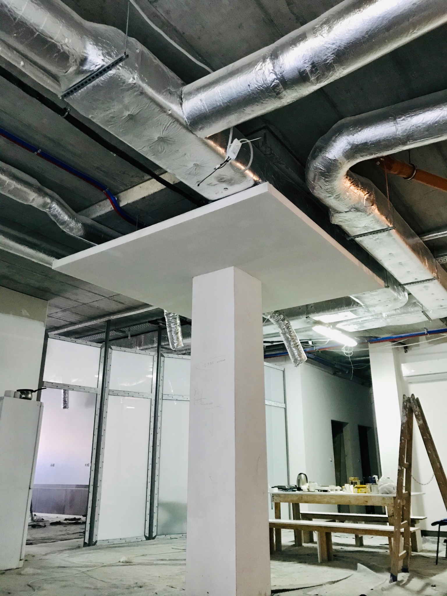 Niedokończona instalacja kanałów wentylacyjnych owiniętych folią aluminiową pod betonowym sufitem w budynku komercyjnym, widoczne elementy konstrukcyjne i tymczasowe oświetlenie.