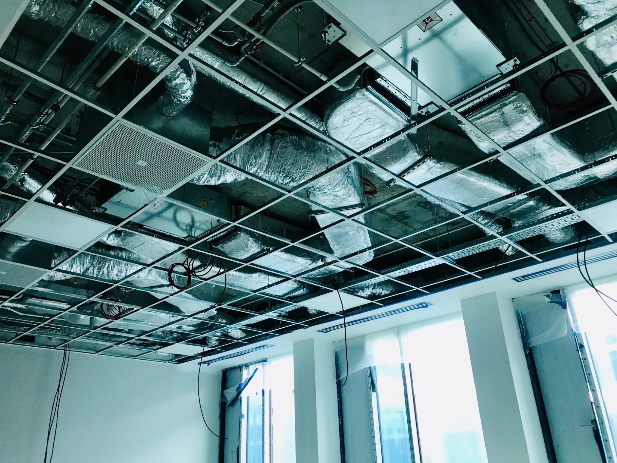 Sufit podwieszany z widocznymi elementami instalacji wentylacyjnej w budynku w Skierniewicach, widoczne kanały wentylacyjne w izolacji, kratka wentylacyjna oraz okablowanie.