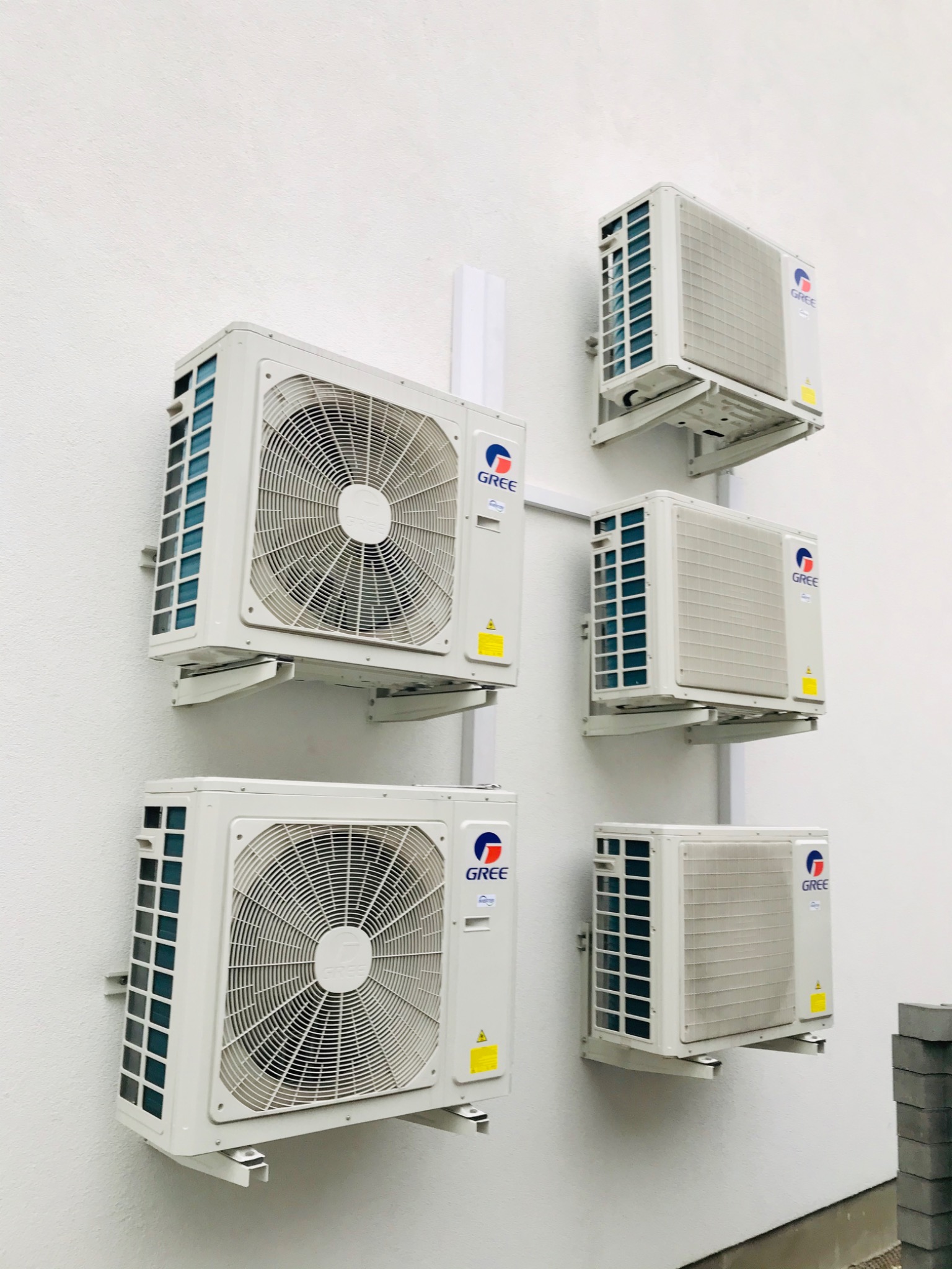 Biała ściana budynku z zamontowanymi, zewnętrznymi jednostkami klimatyzacji marki Gree, rozmieszczonymi w dwóch kolumnach i połączonymi białymi rurami.