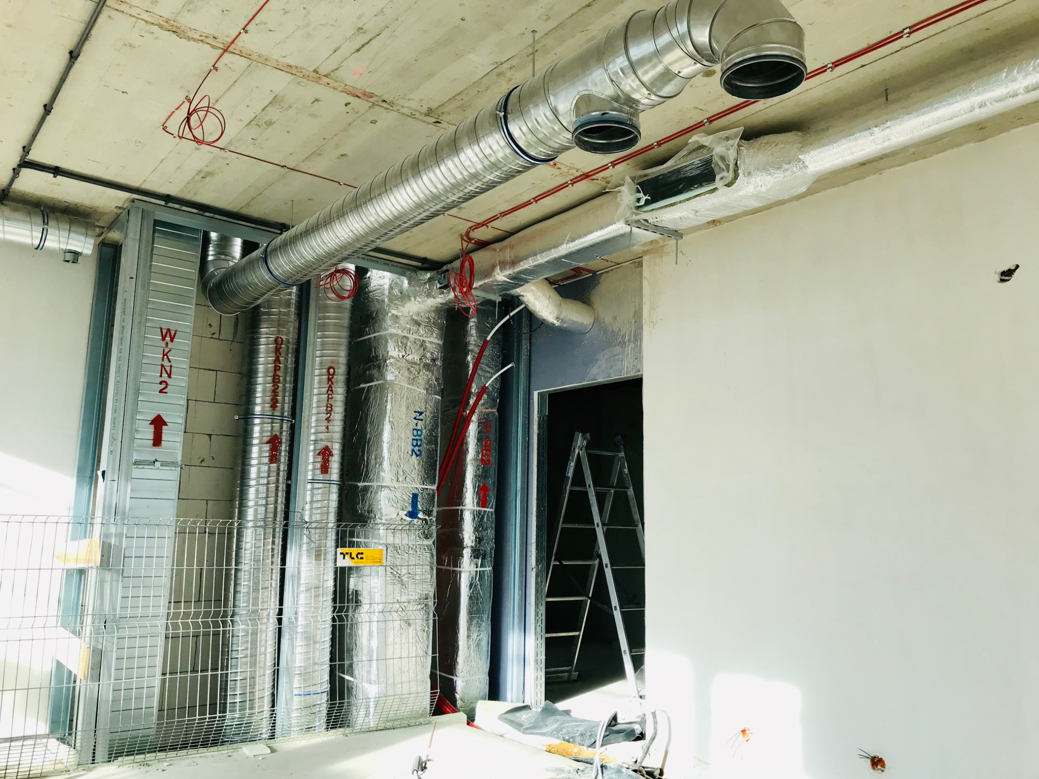 Wnętrze w trakcie montażu systemu wentylacyjnego: widoczne metalowe rury wentylacyjne pod sufitem, pionowe kanały owinięte folią izolacyjną, drabina oparta o ścianę w przejściu, oraz przewody...