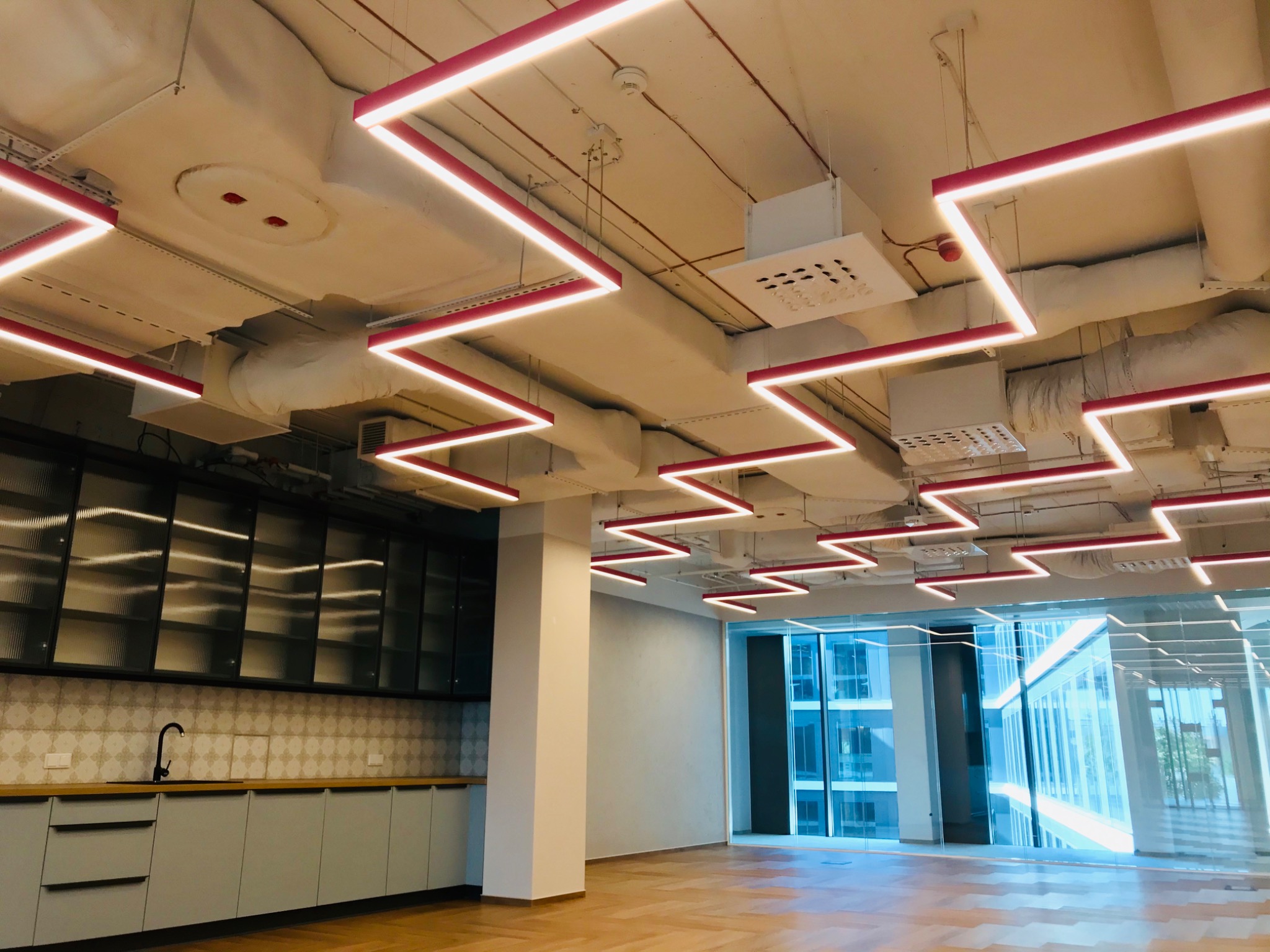 Wnętrze biurowe z widoczną instalacją wentylacyjną pod sufitem i liniowymi lampami LED w kształcie zygzaków, jasna podłoga z drewna, okna od podłogi do sufitu, zabudowa kuchenna z szafkami...
