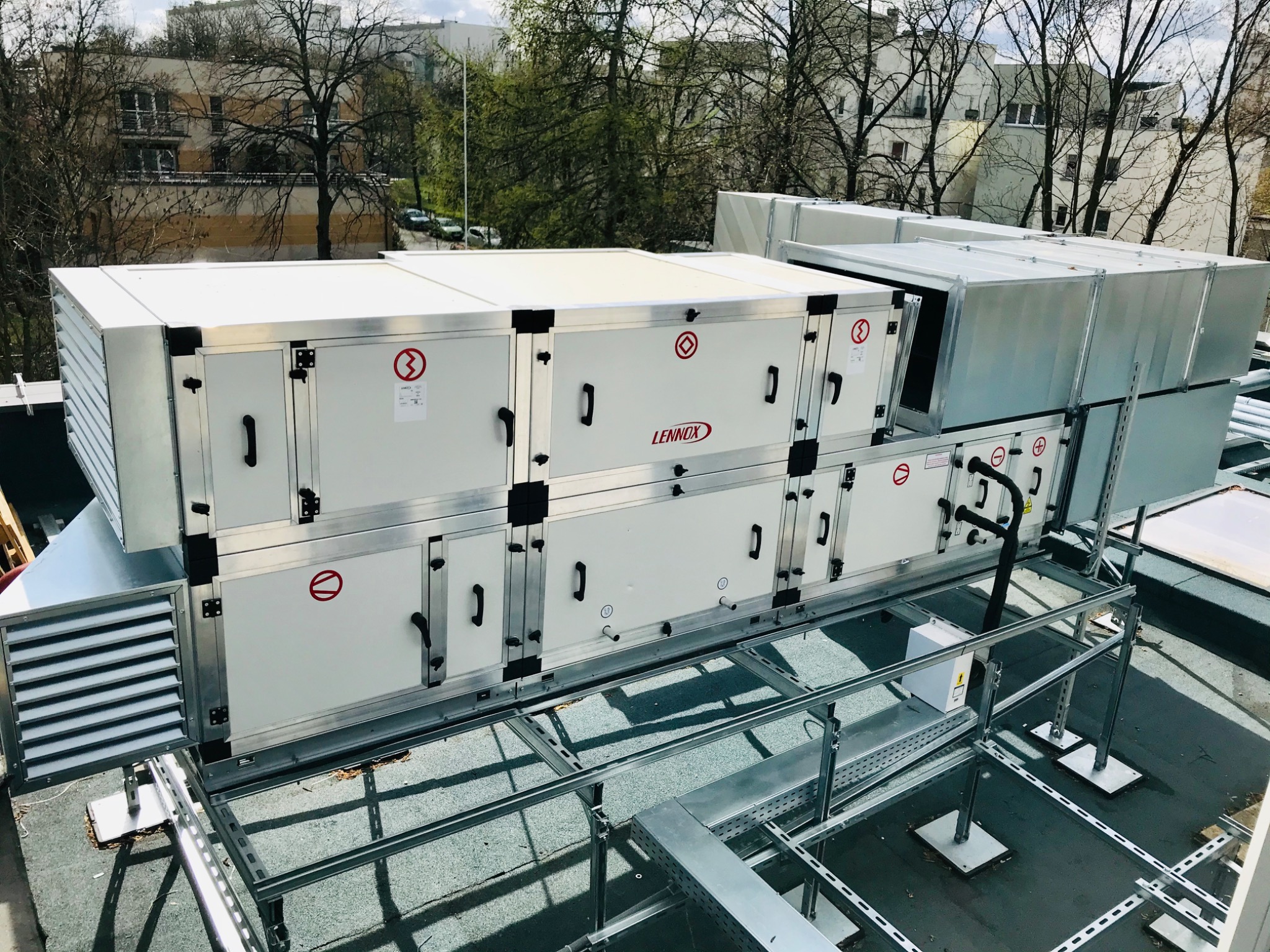 Zewnętrzna instalacja centrali wentylacyjnej Lennox na dachu budynku, widoczne moduły z czarnymi uchwytami i czerwonymi symbolami, osadzone na stalowej konstrukcji wsporczej, w tle drzewa i budynki.