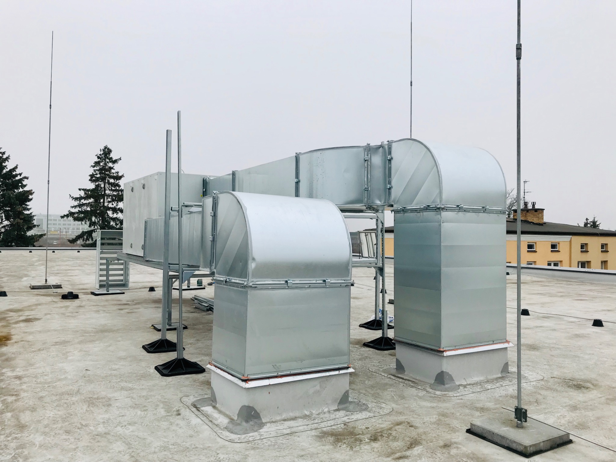 System kanałów wentylacyjnych z ocynkowanej blachy na dachu budynku, z widocznymi elementami mocującymi i odgromnikami, na tle pochmurnego nieba.