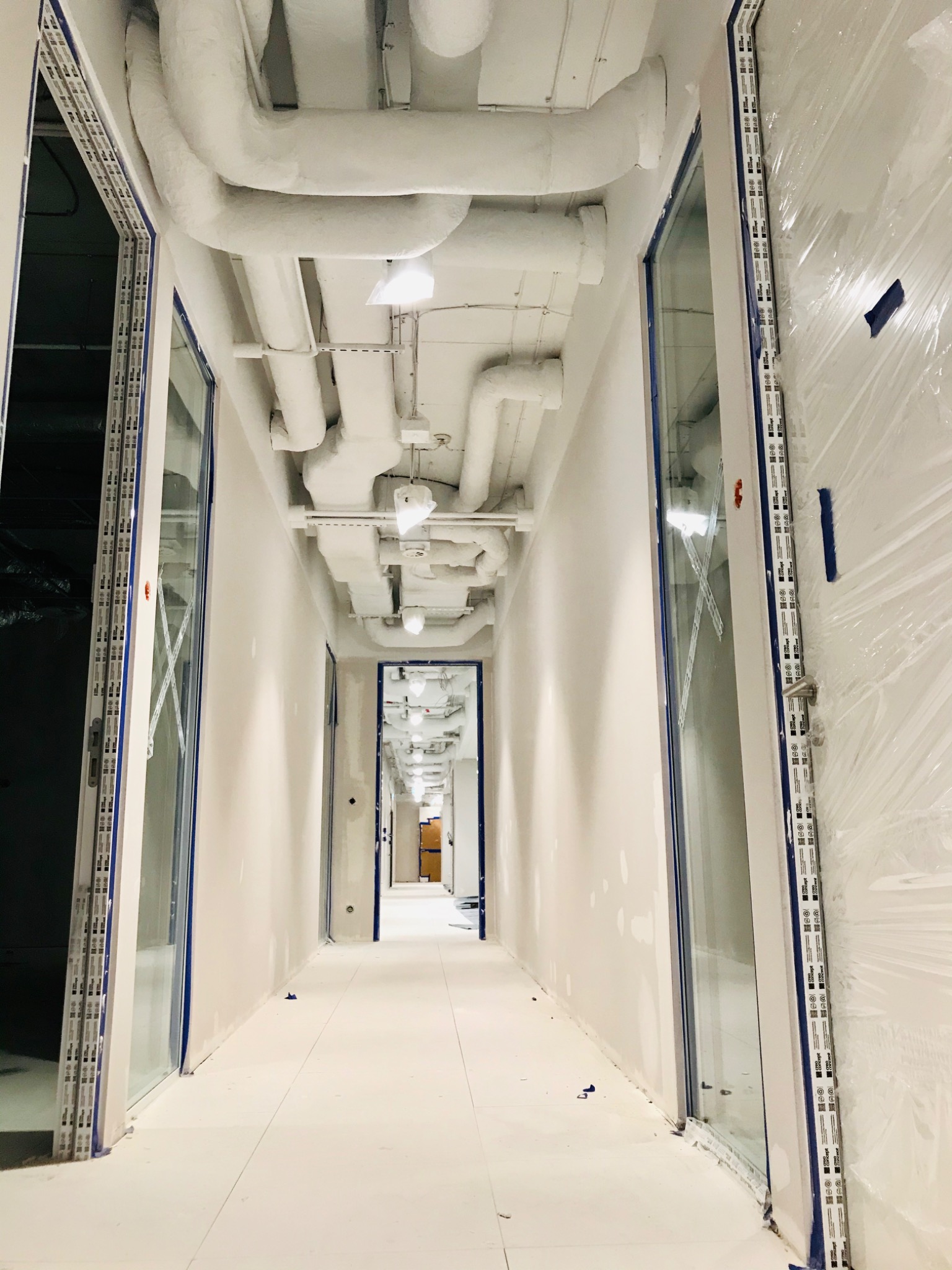 Długi korytarz w budynku komercyjnym w trakcie prac wykończeniowych, widoczne białe rury wentylacyjne pod sufitem, zabezpieczone folią okna i ściany, niebieska taśma malarska.