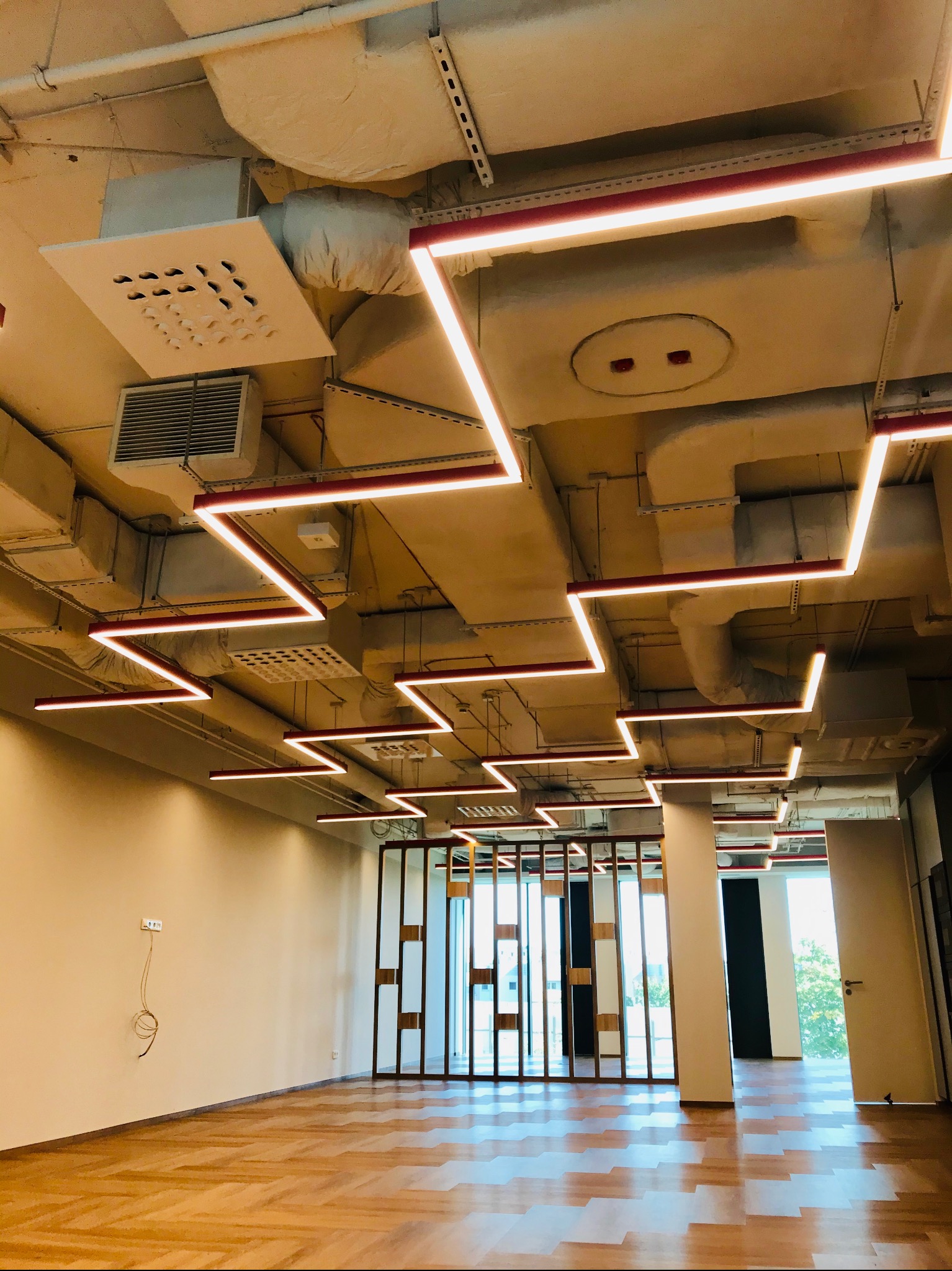 Wnętrze biurowe z widocznymi elementami wentylacji i klimatyzacji pod sufitem, w tym kanały wentylacyjne, kratki nawiewne i system oświetlenia liniowego LED w kształcie zygzaka.